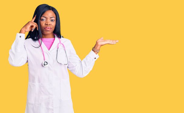 Jonge Afro-Amerikaanse vrouw draagt dokter stethoscoop verward en geïrriteerd met open palm met kopieerruimte en wijzende vinger naar het voorhoofd. Denk er eens over na..  - Foto, afbeelding
