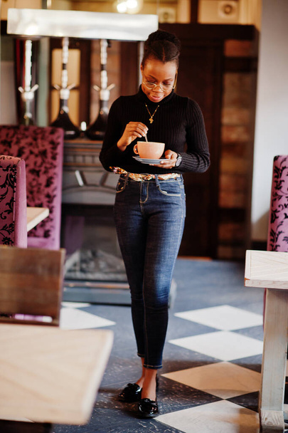 African woman in black sweater and eyeglasses posed at cafe with cup of hot drink. - 写真・画像
