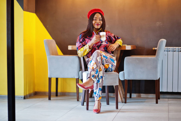 Enthusiastic african american woman in trendy coloured outfit with red beret chilling in cozy cafe with cup of hot latte in hands. - Fotografie, Obrázek