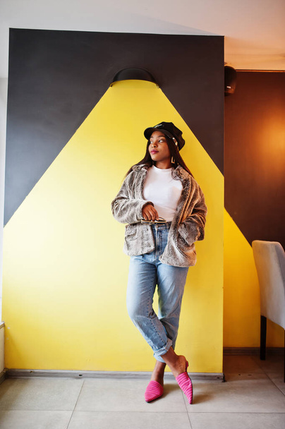 Fashionable african american woman in black cap chilling in cozy cafe and fur against yellow wall. - Foto, imagen