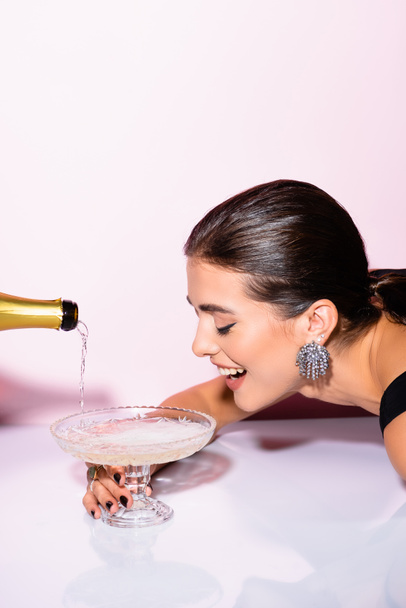 mujer alegre mirando el champán en el vaso cerca de la botella sobre blanco - Foto, imagen