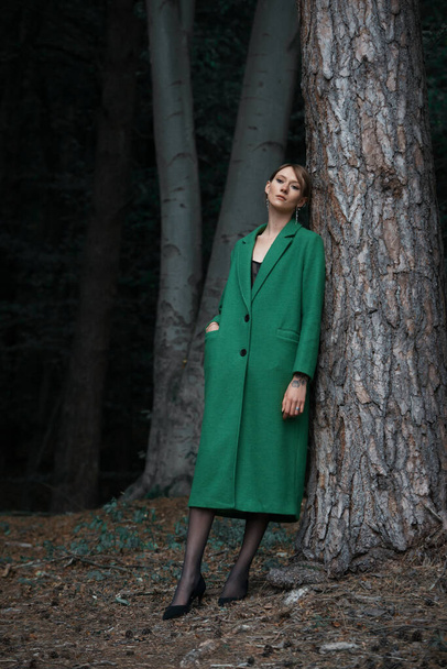 Модная молодая женщина в зеленом пальто позирует в лесу рядом с деревом - Фото, изображение