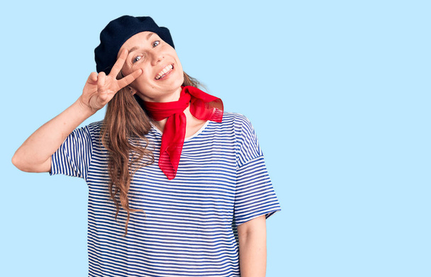 Joven hermosa mujer rubia con boina francesa y camiseta a rayas haciendo símbolo de paz con los dedos sobre la cara, sonriendo alegre mostrando la victoria  - Foto, imagen