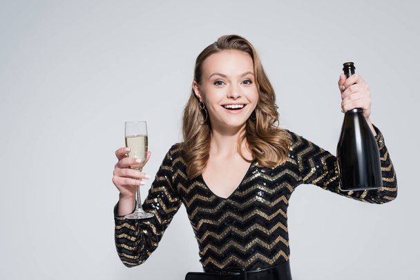 fröhliche Frau hält Glas Champagner und Flasche isoliert auf grau - Foto, Bild