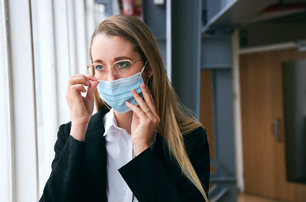健康の流行の間に現代オフィスに立つマスクを身に着けている若いビジネスマンの女性 - 写真・画像