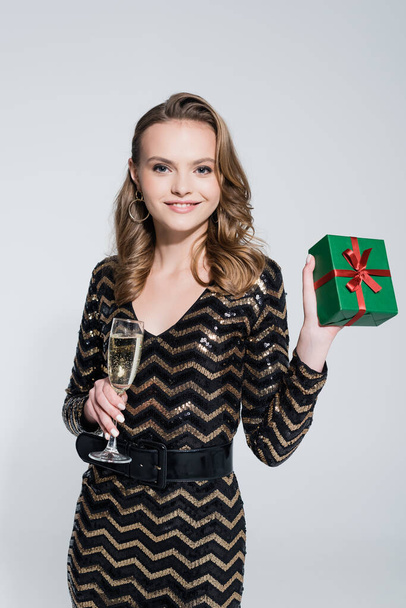 boldog fiatal nő kezében pohár pezsgő és csomagolt karácsonyi ajándék elszigetelt szürke  - Fotó, kép