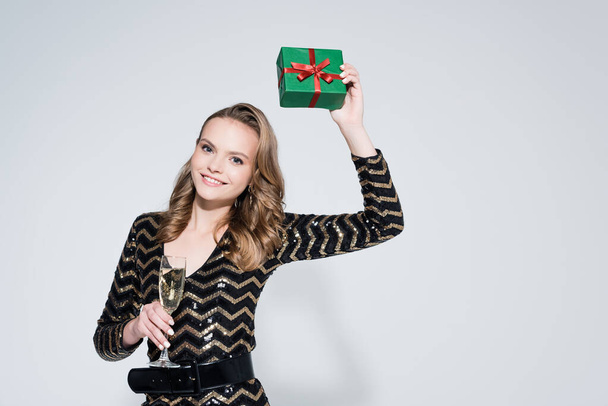 cheerful young woman holding glass of champagne and wrapped christmas present on grey  - Foto, imagen