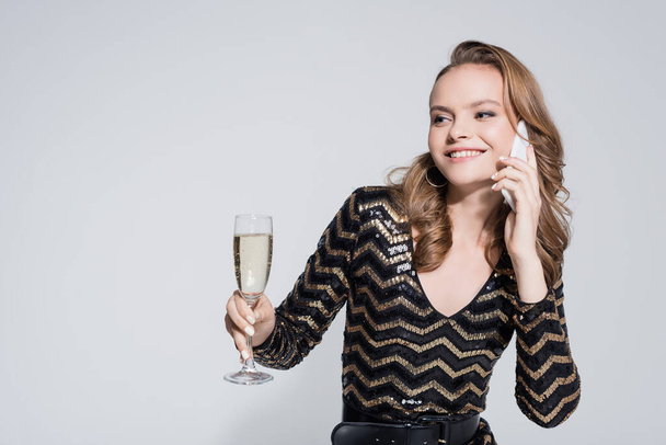 uśmiechnięta młoda kobieta trzymająca kieliszek szampana i rozmawiająca na smartfonie odizolowana na szaro  - Zdjęcie, obraz