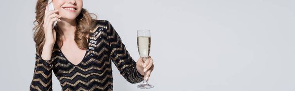 przycięty widok uśmiechniętej kobiety trzymającej kieliszek szampana i rozmawiającej na smartfonie odizolowanym na szarym, banerze - Zdjęcie, obraz
