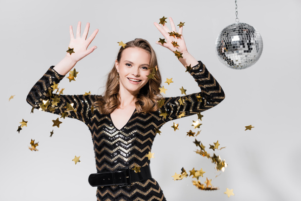 cheerful young woman standing near disco ball and falling confetti on grey - Φωτογραφία, εικόνα