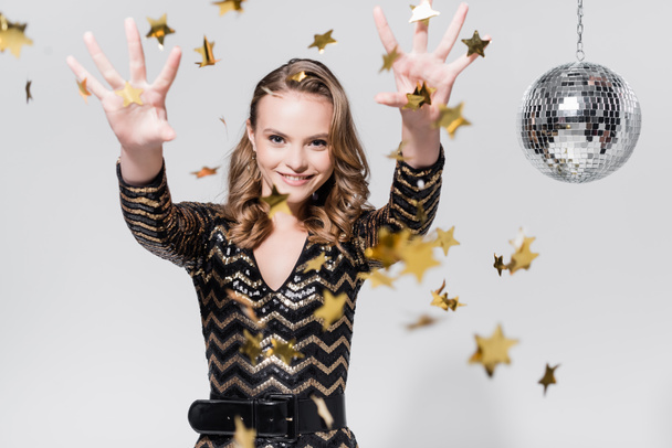 happy young woman standing near disco ball and throwing confetti on grey - Photo, image
