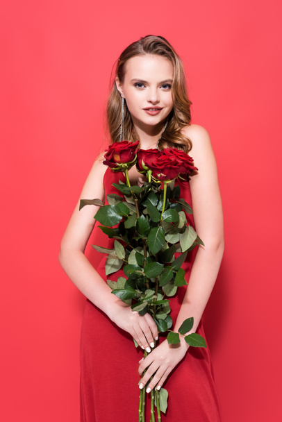 happy young woman holding roses and looking at camera on red  - Фото, зображення