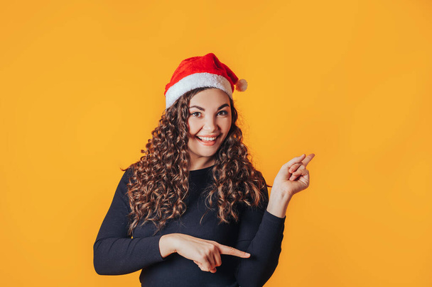 黒のジャケットに巻き毛の美しい女性。空の空間への側面への額のジェスチャー。赤いサンタの帽子。クリスマスや新年のための広告バナーのための写真. - 写真・画像