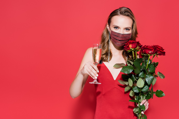 young woman in protective mask with rhinestones holding glass of champagne and roses on red  - Photo, Image