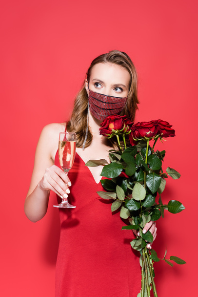 young woman in protective mask with rhinestones holding roses and glass of champagne on red  - Zdjęcie, obraz