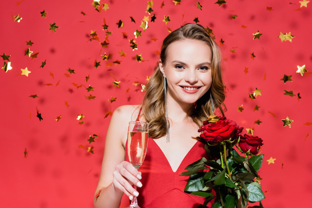 femme heureuse tenant des roses et un verre de champagne près de tomber confettis sur rouge - Photo, image