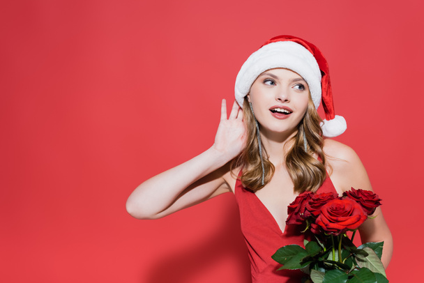 feliz joven en sombrero de Santa sosteniendo rosas y escuchando en rojo  - Foto, Imagen