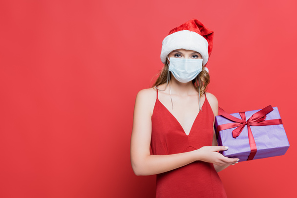 junge Frau mit Weihnachtsmütze und medizinischer Maske hält Weihnachtsgeschenkbox auf rot - Foto, Bild