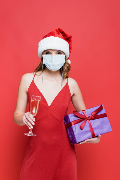 junge Frau mit Weihnachtsmütze und medizinischer Maske mit Weihnachtsgeschenk und einem Glas Champagner auf Rot - Foto, Bild
