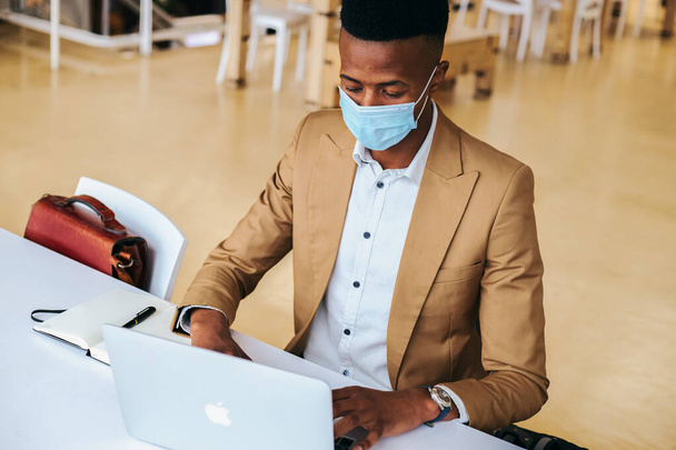 Junger Geschäftsmann mit Maske arbeitet während Gesundheitspandemie am Laptop am heißen Schreibtisch im Büro - Foto, Bild