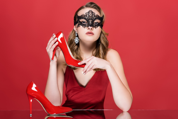 jeune femme élégante en noir masque de carnaval tenant chaussure sur rouge - Photo, image
