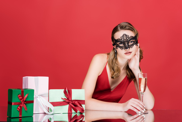 jeune femme en élégant masque de dentelle tenant verre de champagne près de cadeaux de Noël sur rouge - Photo, image