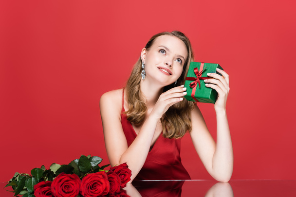 young dreamy woman looking up near roses while holding christmas gift box on red - 写真・画像