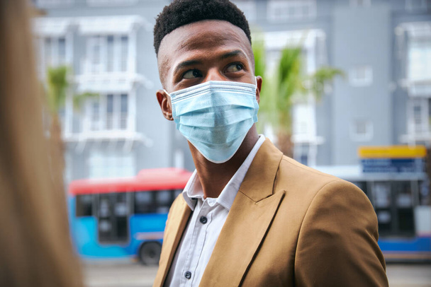 Jonge zakenman in masker die buiten het moderne kantoorgebouw staat tijdens de gezondheidspandemie - Foto, afbeelding