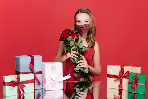 woman in protective mask with rhinestones holding roses near christmas presents on red - Photo, Image