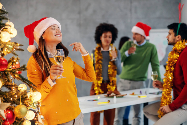 サンタさんの帽子をかぶった若い笑顔の実業家がシャンパンを持ち、クリスマスツリーの横に立って新年の前夜を祝います。背景には彼女の同僚もいます. - 写真・画像