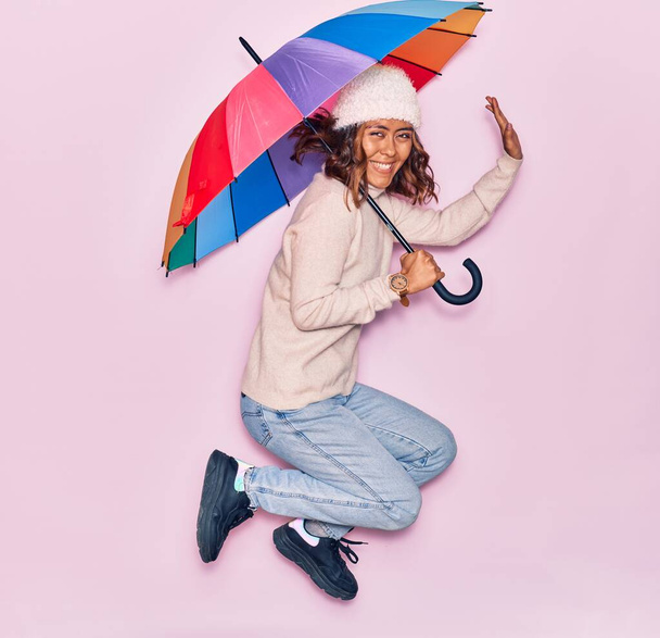 Fiatal gyönyörű latin nő kezében esernyő mosolyogva boldog. Ugrás mosollyal az arcon át elszigetelt rózsaszín háttér - Fotó, kép