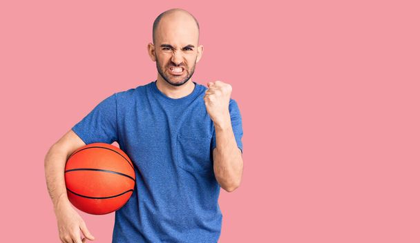 Joven hombre guapo sosteniendo pelota de baloncesto molesto y frustrado gritando con ira, gritando loco de ira y la mano levantada  - Foto, Imagen