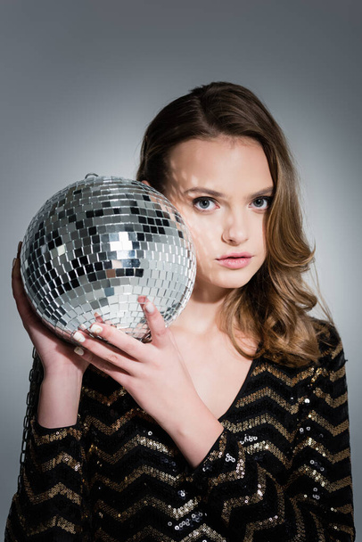 young woman looking at camera while holding disco ball on grey - Фото, зображення