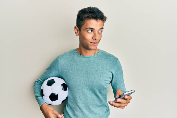 Νεαρός όμορφος Αφροαμερικάνος που κρατάει μπάλα ποδοσφαίρου κοιτάζοντας το smartphone χαμογελώντας κοιτώντας στο πλάι και κοιτάζοντας αλλού σκεπτόμενος.  - Φωτογραφία, εικόνα