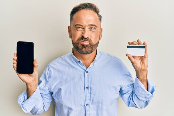 Handsome middle age man holding smartphone and credit card puffing cheeks with funny face. mouth inflated with air, catching air.  - Fotoğraf, Görsel