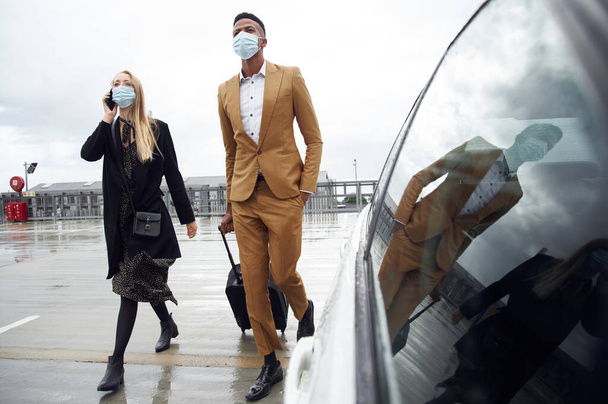 Pareja de negocios con máscaras fuera del aeropuerto para viajes de negocios con equipaje durante la pandemia de salud - Foto, imagen