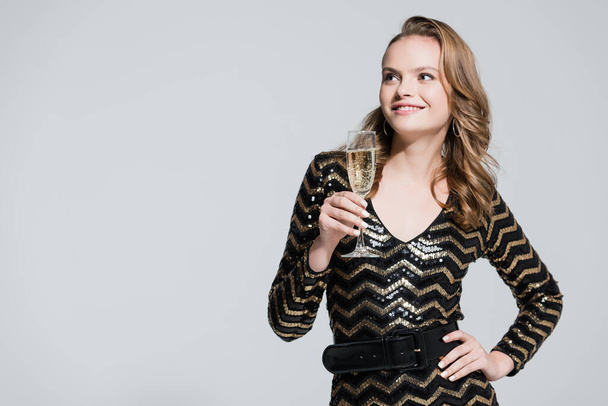 cheerful woman holding glass of champagne and standing with hand on hip isolated on grey - Photo, Image