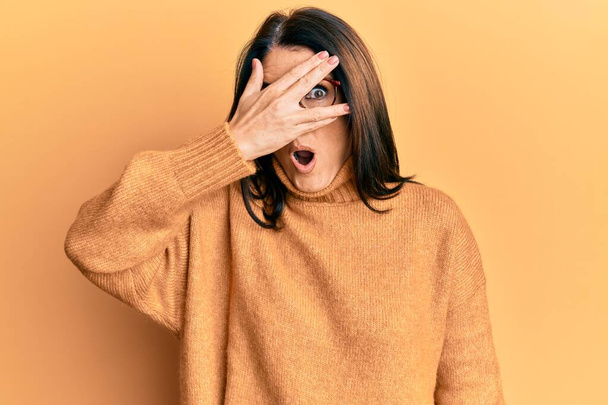 Middelbare leeftijd brunette vrouw dragen casual winter trui over gele achtergrond gluren in shock bedekking gezicht en ogen met de hand, kijken door vingers bang  - Foto, afbeelding