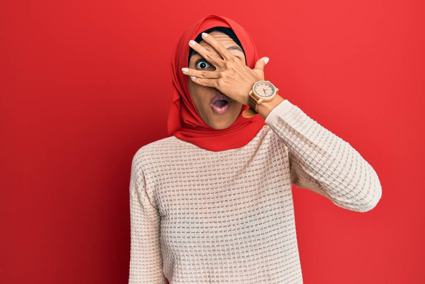 Młoda Afroamerykanka w tradycyjnym islamskim hidżabskim szaliku, zaglądająca ręką w szok zakrywający twarz i oczy, patrząca przez palce.  - Zdjęcie, obraz