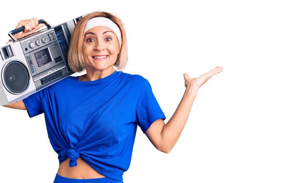 Young blonde woman wearing sportswear holding boombox, listening to music celebrating victory with happy smile and winner expression with raised hands  - Fotografie, Obrázek