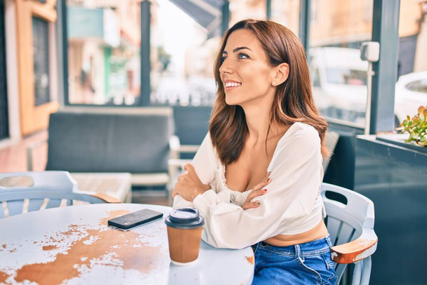 Joven mujer hispana sonriendo feliz sentada en la terraza de la cafetería. - Foto, imagen