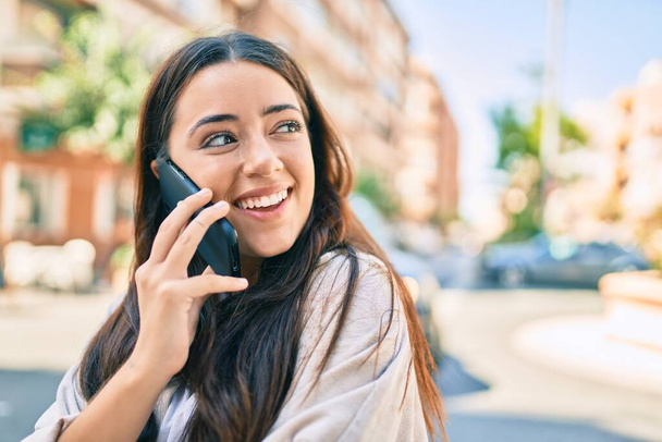 Молодая латиноамериканка улыбается счастливо разговаривая по смартфону в городе. - Фото, изображение