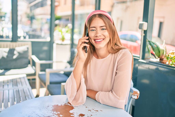 Νεαρή καυκάσια κοπέλα χαμογελά ευτυχισμένη μιλώντας στο smartphone στη βεράντα του καφενείου. - Φωτογραφία, εικόνα