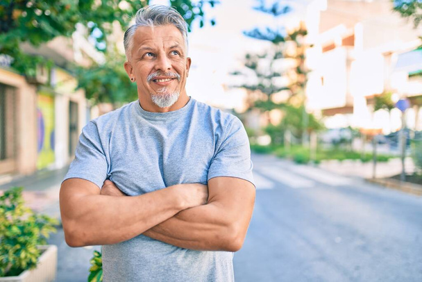 Hombre hispano de pelo gris de mediana edad con los brazos cruzados sonriendo feliz en la ciudad. - Foto, imagen