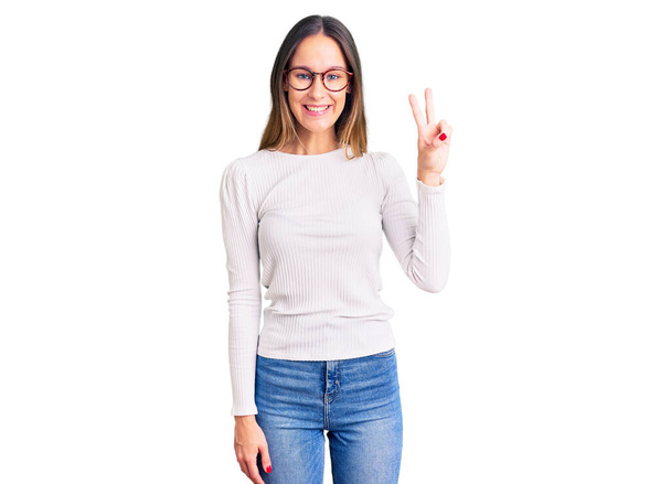 Beautiful brunette young woman wearing casual white sweater and glasses showing and pointing up with fingers number two while smiling confident and happy.  - Photo, image