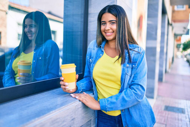 Joven latina sonriendo feliz bebiendo llevar café a la ciudad. - Foto, Imagen