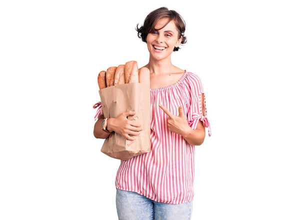 Mooie jonge vrouw met kort haar vasthouden papieren zak met brood lachend gelukkig wijzend met hand en vinger  - Foto, afbeelding
