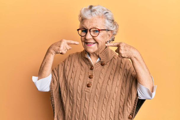 Donna anziana dai capelli grigi indossa abiti casual e occhiali sorridenti allegri mostrando e indicando con i denti e la bocca le dita. concetto di salute dentale.  - Foto, immagini