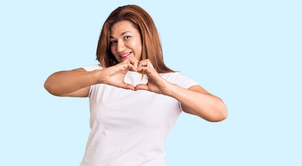 Mulher latina de meia idade vestindo camisa branca casual sorrindo no amor fazendo forma de símbolo do coração com as mãos. conceito romântico.  - Foto, Imagem