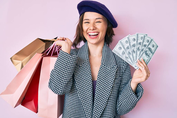 Mladá krásná žena v baretu drží nákupní tašky a dolary bankovky s úsměvem a hlasitě se směje, protože legrační bláznivý vtip.  - Fotografie, Obrázek